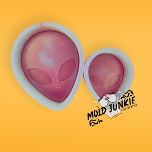 May include: Two silicone molds in the shape of alien heads. The molds are white with a pink and purple shimmer. The larger mold is on the left and the smaller mold is on the right. The text "Mold Junkie Feel the Fever" is in the lower right corner of the image.
