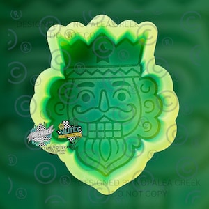 May include: A green silicone mold featuring a nutcracker face design with a crown, mustache, and beard. The mold has a scalloped edge and includes the text Silitex Get Creative Get Sil and Nopalea Creek A Family of Brands Sister Companies Shared Vision.