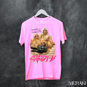 May include: A pink t-shirt featuring a graphic of Cardi B and the text "BAD BITCHES HAVE FUN, HATH HOES MAKE EXCUSES" and "CARDI B" in a bold, stylized font. The graphic shows three women in fur coats.