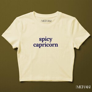 Peut inclure: Un haut court à manches courtes de couleur crème avec le texte bleu foncé "spicy capricorn" imprimé sur la poitrine. L'étiquette intérieure du col affiche le nom de la marque "MERAKI" dans une police simple, assortie à la marque en bas.