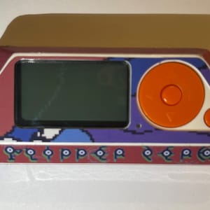 May include: A retro handheld gaming device with a rectangular screen and an orange control pad. The device has a red and white border with pixelated blue and purple accents. The bottom edge features stylized text.