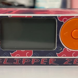 Puede incluir: Un dispositivo electrónico de mano blanco y naranja con una pantalla negra. El dispositivo tiene un botón circular con flechas direccionales y un botón más pequeño. El dispositivo tiene un patrón rojo y azul con el texto "FLIPPER ZERO" en la parte inferior.
