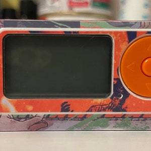 Puede incluir: Un dispositivo electrónico de mano blanco y naranja con un diseño gráfico colorido. El dispositivo tiene una pantalla, un botón circular con flechas direccionales y un botón más pequeño.