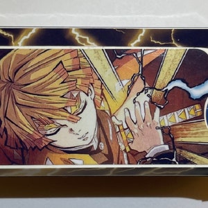Könnte beinhalten: Ein weißes Plastikgehäuse mit einem schwarzen und gelben Blitz-Muster. Das Gehäuse zeigt einen farbenfrohen Anime-Charakter mit orangefarbener und gelber Kleidung. Die Augen des Charakters sind geschlossen und er hält ein Schwert. Das Gehäuse hat ein kleines rundes Bild des Charakters in der unteren rechten Ecke.