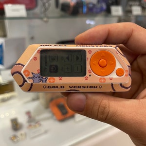Könnte beinhalten: Eine handgehaltene Videospielkonsole in tan und orange. Die Konsole hat einen kleinen Bildschirm, Knöpfe und ein Steuerkreuz. Die Konsole ist mit "Pocket Monsters Gold Version" beschriftet.