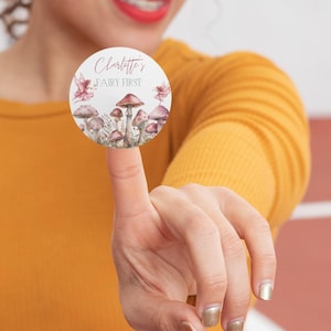 May include: A round sticker with a watercolor illustration of mushrooms and fairies. The text on the sticker reads "Charlotte's Fairy First".