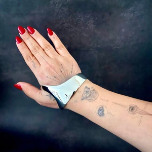 May include: A silver metal cuff bracelet with a curved, open design. The bracelet is worn on a person's wrist.