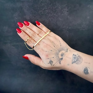 Puede incluir: Conjunto de anillos de oro con una banda fina y una cadena que conecta un anillo al dedo medio. El conjunto de anillos se lleva en una mano con esmalte de uñas rojo.