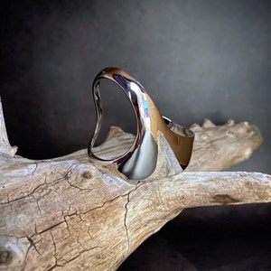 May include: A silver, abstract, organic-shaped ring with a smooth, polished finish. The ring is displayed on a piece of driftwood.