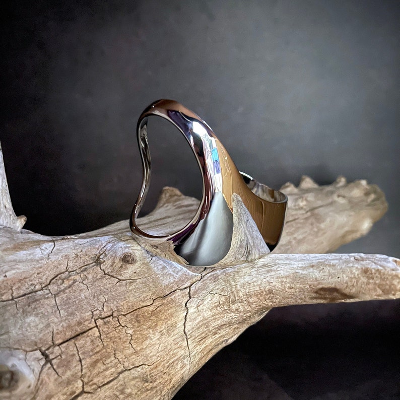 May include: A silver, abstract, organic-shaped ring with a smooth, polished finish. The ring is displayed on a piece of driftwood.