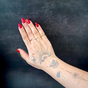 May include: A gold-colored bar ring worn on a hand with red nail polish. The hand has several tattoos, including a sunburst design and celestial imagery. The background is a dark, textured surface.