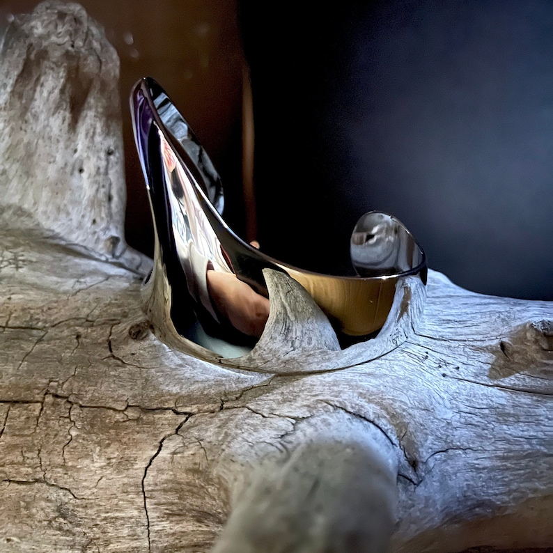 May include: A silver metal cuff bracelet with a curved, organic shape. The bracelet is resting on a piece of driftwood.
