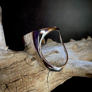 May include: A silver, abstract, open-ring style bracelet with a smooth, polished finish. The ring is resting on a piece of weathered driftwood.