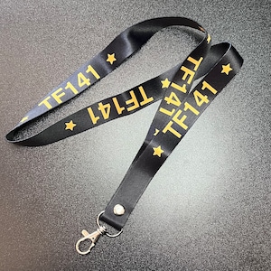 Puede incluir: Lanyard negro con un clip dorado y el texto "TF141" repetido en ambos lados. El texto está en negrita, sin serif.