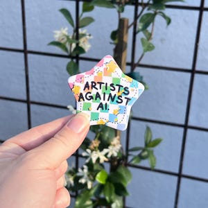 Puede incluir: Un pin con forma de estrella que presenta un patrón de cuadros en tonos pastel rosa, verde y azul. El texto "ARTISTS AGAINST AI." está impreso en letras negras, negritas y sans-serif en el centro, acentuado por pequeños iconos de estrellas amarillas.