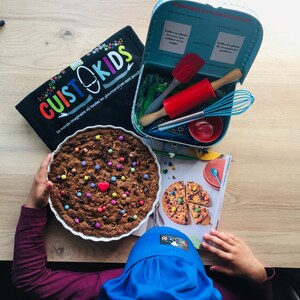 以下が含まれることがあります： 青いシェフの帽子をかぶった子供が、カラフルなキャンディーで飾られたクッキーを持っています。クッキーは白い陶器の皿に入っています。青い白のトランクがテーブルの上に開いており、中にはキッチンツールが入っています。トランクには「Bienvenue au Club Cuisito Kids!」という文字が書かれています。テーブルの上には、クッキーの写真が載っている料理本が開かれています。