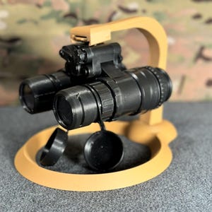 May include: A black night vision scope with a tan plastic stand. The scope has a lens cover on each side.