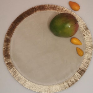 May include: A round beige placemat with a gold fringe border. A green mango and two small yellow fruits are on the placemat.