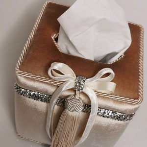 May include: A beige velvet tissue box cover with a white tissue inside. The cover has a white ribbon with a rhinestone bow and a beige tassel.