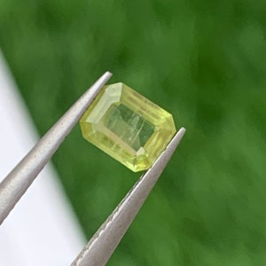 May include: A light green gemstone with a rectangular shape, held by tweezers against a green background.