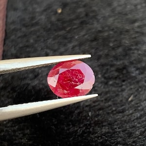 Może przedstawiać: Czerwony kamień szlachetny o okrągłym szlifie, trzymany w pęsecie na czarnym tle.