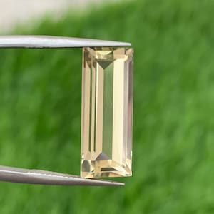 May include: A light yellow faceted gemstone in a rectangular shape, held by tweezers against a green background.