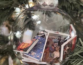 May include: Clear glass Christmas ornament filled with miniature book pages. The ornament hangs from a silver string on a green Christmas tree. The miniature books have colorful covers and the word "Shakespeare" is visible.