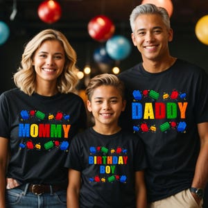 May include: A family of three wearing black t-shirts with colorful Lego block designs. The shirts say "Mommy", "Daddy", and "Birthday Boy".