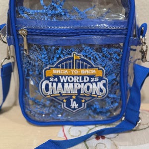 May include: Clear crossbody bag with blue trim and a blue shoulder strap. The bag features a zipper closure and a graphic that reads "Back-to-Back 24 World 25 Champions" with a team logo. The bag is filled with blue crinkle paper.