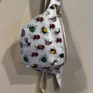 May include: White fanny pack with a zipper closure and a gold zipper pull. The fanny pack has a white background with a repeated pattern of various Avengers characters, including Iron Man, Captain America, Thor, and Hulk.
