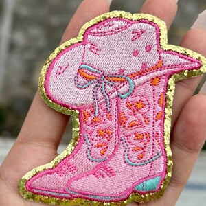 May include: Pink embroidered cowboy boot and hat patch with gold glitter edging. The patch features orange and teal accents on the boot and a bow on the hat. This decorative patch is ideal for adding a touch of Western flair to clothing or accessories.