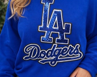 May include: A royal blue crewneck sweatshirt featuring a large, sequined 'LA' logo in blue with a white and black border, positioned above the word 'Dodgers' in a white and black script font, also embellished with blue sequins.