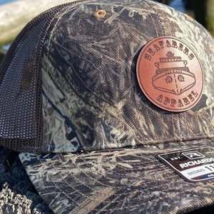 May include: A brown and black camouflage trucker hat with a brown leather patch that says "Seafarers Apparel" and has a ship logo. The hat is sitting on a wooden post.