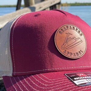 May include: A maroon and beige trucker hat with a brown leather patch that says "Seafarred Apparel" and a jet ski graphic. The hat is sitting on a wooden surface.