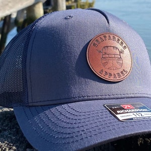 Puede incluir: Una gorra de camionero de malla azul y negra con un parche de cuero marrón que dice "Sentinels Apparel" con un gráfico de una persona con casco. La gorra está sobre una superficie de madera.