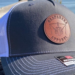 Könnte beinhalten: Eine graue und weiße Trucker-Kappe mit einem braunen Leder-Patch, auf dem "Sailfarers Apparel" mit einem Schiffsdesign steht. Die Kappe ist von Richardson 112.