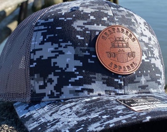 May include: A black and white camouflage trucker hat with a brown leather patch that says "Seafarers Apparel" and a graphic of a boat.