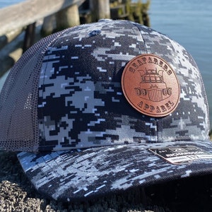 Könnte beinhalten: Eine schwarze und weiße Camouflage-Trucker-Kappe mit einem braunen Leder-Patch, auf dem "Seafarers Apparel" und eine Grafik eines Bootes steht.