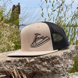 Könnte beinhalten: Eine beige und schwarze Baseballkappe mit einem schwarzen Jetski-Motiv, das auf der Vorderseite gestickt ist. Die Kappe hat ein Netzrücken.