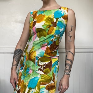 Op de afbeelding: Een mouwloze vintage jurk met een levendig bloemenpatroon. De jurk heeft een ronde hals, twee voorzakken en een kleurrijke print met tinten blauw, groen, geel en bruin. De zakken zijn roze afgewerkt.