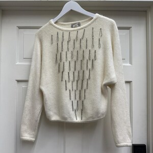 May include: Cream-colored sweater with long sleeves and a crew neck. The sweater features a pattern of vertical, dark gray embellishments. The garment is hanging on a white hanger against a white door.