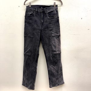 May include: Dark gray denim jeans with a high waist and straight leg. The jeans feature a unique design with rectangular pockets on the thighs and knees, and vertical seams down the front and back of the legs. The jeans are hanging on a metal hanger.