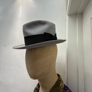 Puede incluir: Un sombrero fedora de color gris claro con una banda ancha de cinta de grosgrain negra atada en un lazo en el lateral. El sombrero tiene una copa estructurada con un pliegue central y un ala plana, sobre un maniquí de tono neutro.
