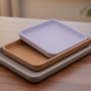 May include: A set of three rectangular trays stacked on top of each other. The top tray is light purple, the middle tray is tan, and the bottom tray is a muted gray. Each tray features raised edges and a smooth, matte finish with rounded corners.