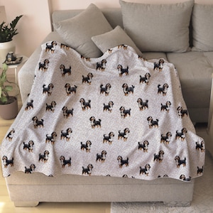 May include: A light gray blanket draped over a sofa, featuring a repeating pattern of small, black, brown, and white dogs. The blanket is made of a soft material and is folded over the sofa cushions.