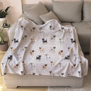 May include: A light gray blanket draped over a sofa, featuring a repeating pattern of pug dog illustrations in various colors. The blanket is made of a soft material and is a cozy home decor item.