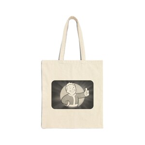 May include: A beige canvas tote bag featuring a rectangular graphic print in the center. The print depicts a cartoon character with a smiling face and a winking eye, giving a thumbs-up gesture against a dark, radiating circular background.