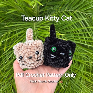 May include: Two crocheted teacup kitty cat amigurumi toys. One is beige with brown eyes and the other is black with green eyes. The text "Pdf Crochet Pattern Only" and "Thick Thumb Crochet" is below the toys.