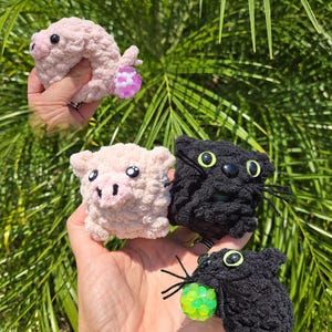 May include: A collection of handmade crochet toys, including a pink pig-shaped donut, a square pink pig, and two black cat-shaped toys. The toys have button eyes and are held in a hand against a backdrop of green foliage.