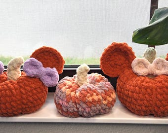 Peut inclure: Trois citrouilles faites à la main au crochet, dans des tons orange, rose et violet. Deux citrouilles ont de grands accents en forme d'oreilles et des nœuds. Les citrouilles sont exposées sur un rebord de fenêtre blanc avec une plante en arrière-plan.